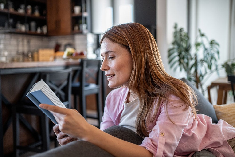Mujer relajándose en un sofá y leyendo un libro durante su recuperación tras una cirugía de corrección de la visión LASIK, PRK o EVO ICL.