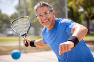 Atractivo hombre mitad hispano de unos 50 años jugando ráquetbol al aire libre en un día soleado, usando gafas deportivas aprobadas por OSHA para proteger su visión mientras vive con tracción vítreomacular, atención brindada por West Texas Eye Associates.