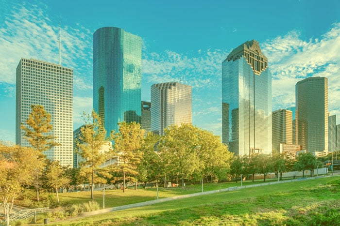 City skyline as seen through cataract-affected vision, showing tall modern skyscrapers with softened outlines, hazy glare, muted colors, and reduced contrast that blur building details and make the bright sky appear diffused.