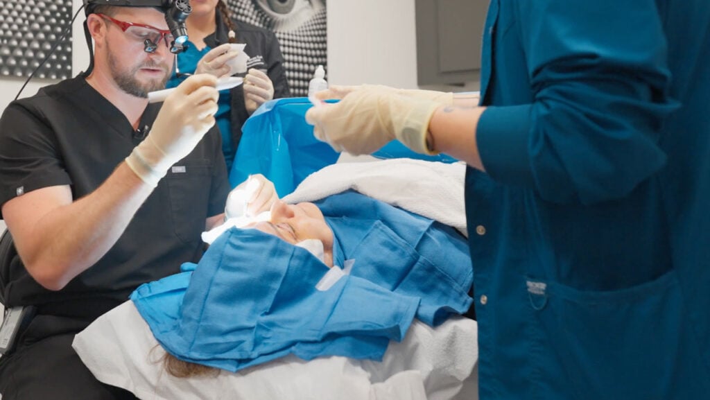 Cirujano oculoplástico realizando una blefaroplastia del párpado inferior a un paciente en un entorno clínico, con campos estériles e instrumental quirúrgico visibles.