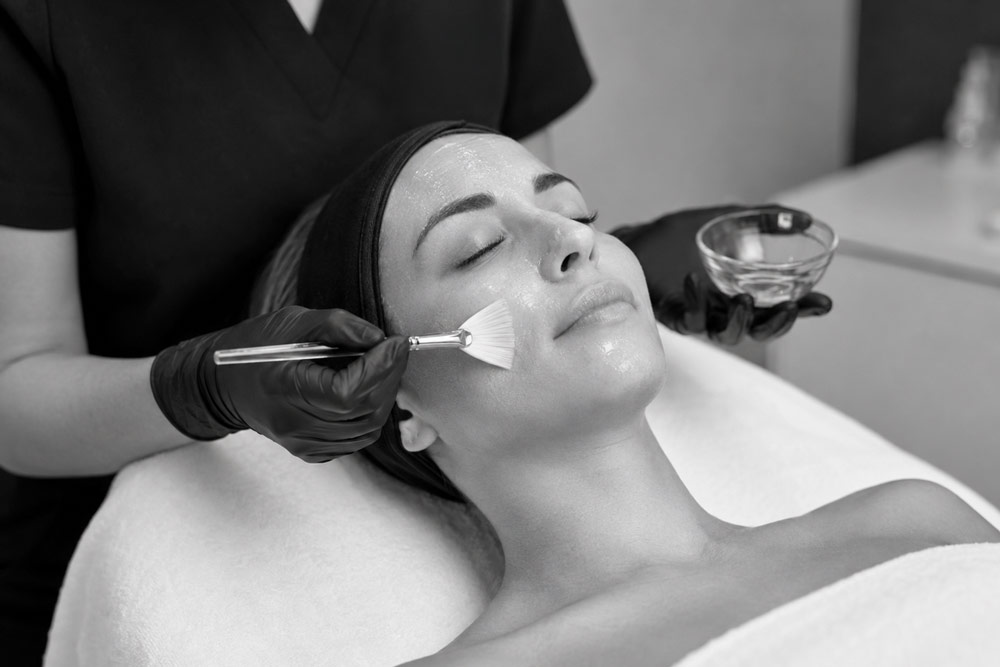 Esthetician applying a chemical peel treatment to a patient’s face in a black-and-white med spa setting at Aesthetic Center of Lubbock.