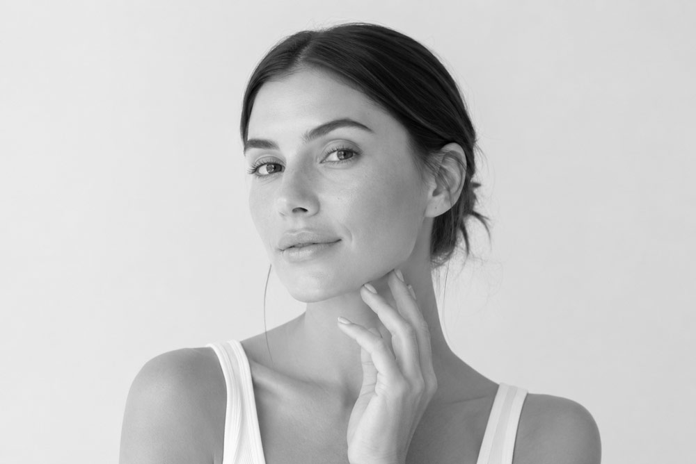Woman with smooth, glowing skin gently touching her jawline after a chin filler treatment in a bright med spa-style portrait.