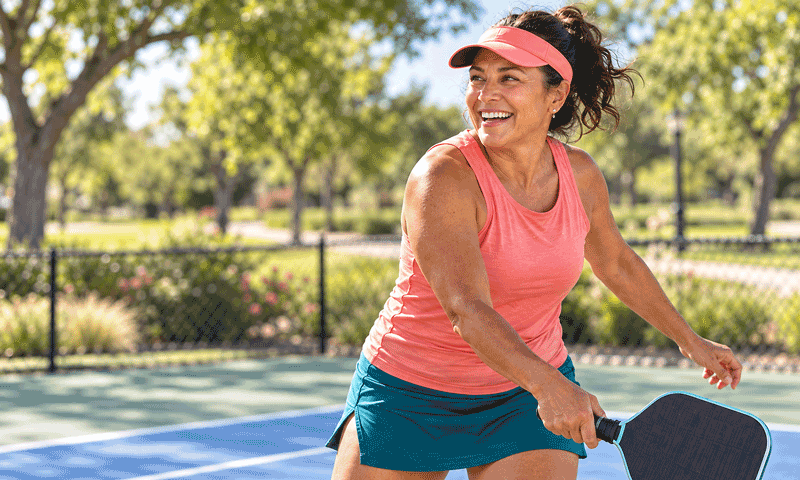 An active 50-year-old woman smiles while playing pickleball on a sunny court surrounded by a green park.