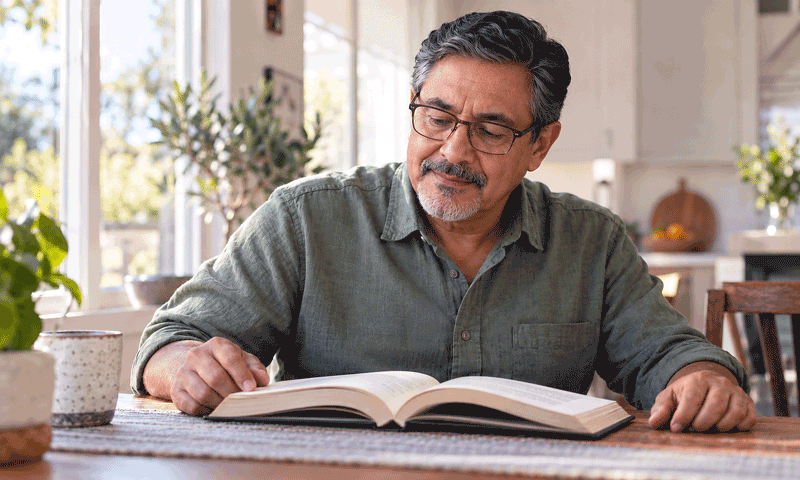 Man wearing reading glasses pauses over an open book at a sunlit kitchen table.